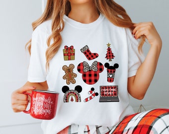Op de afbeelding: Wit T-shirt met een rood-zwart buffalo plaid-patroon met verschillende kerstthema-afbeeldingen, waaronder een peperkoekman, een kerstboom, een kous, een cadeaudoos, een koffiebeker, een snoepstok, een laptop met de tekst "Christmas Movies" en een Mickey Mouse-hoofd met een strik.