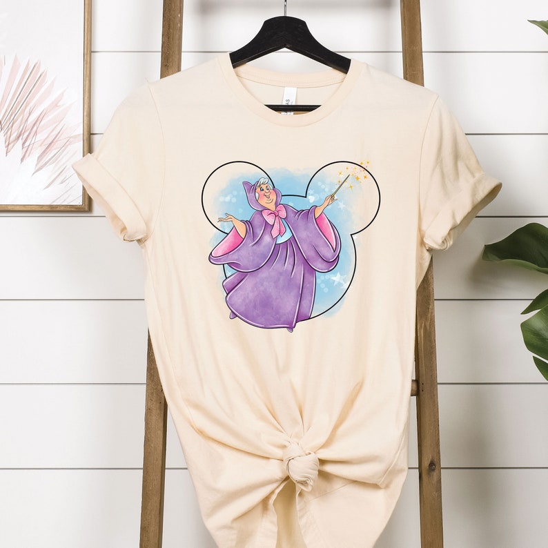 May include: A light beige t-shirt with a cartoon image of the Fairy Godmother from Cinderella. The Fairy Godmother is wearing a purple gown and holding a magic wand. The image is surrounded by a blue and white Mickey Mouse head.