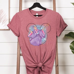 May include: A pink heather t-shirt with a graphic of a fairy godmother in a purple dress with a pink bow, holding a wand with stars, inside a Mickey Mouse head silhouette.