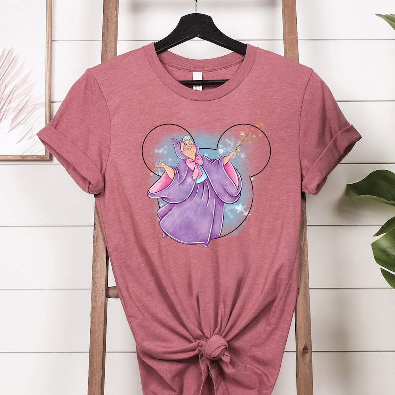 May include: A pink heather t-shirt with a graphic of a fairy godmother in a purple dress with a pink bow, holding a wand with stars, inside a Mickey Mouse head silhouette.