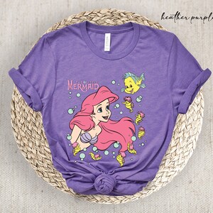 Peut inclure: Un t-shirt en jersey violet chin&eacute; avec un motif d'Ariel de La Petite Sir&egrave;ne. Le motif comprend Ariel, Polochon et S&eacute;bastien. Le texte "La Petite Sir&egrave;ne" est imprim&eacute; au-dessus d'Ariel.