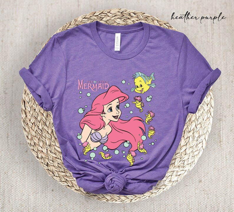 Peut inclure: Un t-shirt en jersey violet chin&eacute; avec un motif d'Ariel de La Petite Sir&egrave;ne. Le motif comprend Ariel, Polochon et S&eacute;bastien. Le texte "La Petite Sir&egrave;ne" est imprim&eacute; au-dessus d'Ariel.