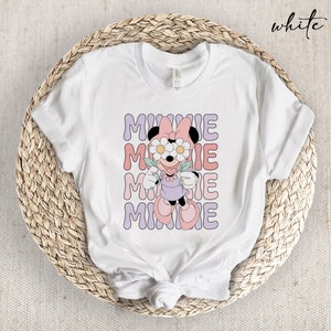 May include: White t-shirt with a graphic of Minnie Mouse wearing flowers over her eyes. The text "MINNIE" is repeated four times in a wavy font.