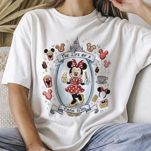 Op de afbeelding: Wit T-shirt met een kleurrijke afbeelding van Minnie Mouse in een rode polkadotjurk, met ijs en een traktatie. Het ontwerp bevat de tekst "The Life of a Park Girl" en park-thema-iconen.