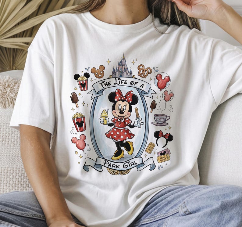 Op de afbeelding: Wit T-shirt met een kleurrijke afbeelding van Minnie Mouse in een rode polkadotjurk, met ijs en een traktatie. Het ontwerp bevat de tekst "The Life of a Park Girl" en park-thema-iconen.