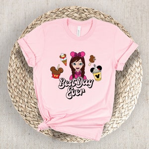 May include: A pale pink t-shirt with a cartoon illustration of a girl with a pink bow, surrounded by ice cream and Mickey Mouse-shaped treats. The text "Best Day Ever" is written in a playful font.