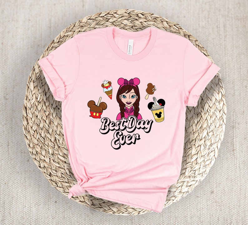 May include: A pale pink t-shirt with a cartoon illustration of a girl with a pink bow, surrounded by ice cream and Mickey Mouse-shaped treats. The text "Best Day Ever" is written in a playful font.