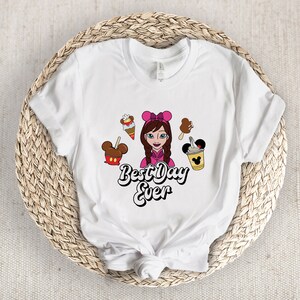 May include: White t-shirt with a cartoon design of a girl with a pink bow and treats. The words "Best Day Ever" are in a fun font. The design includes ice cream, Mickey Mouse-shaped treats, and a drink, perfect for a fun day out.