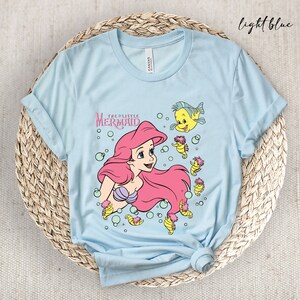 Peut inclure: T-shirt bleu clair avec un motif d'Ariel de La Petite Sir&egrave;ne, entour&eacute;e d'hippocampes et de bulles. Le texte "The Little Mermaid" est imprim&eacute; au-dessus d'Ariel.