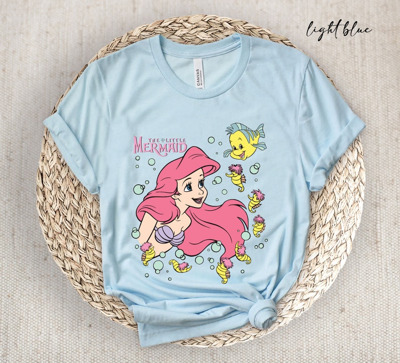 Peut inclure: T-shirt bleu clair avec un motif d'Ariel de La Petite Sir&egrave;ne, entour&eacute;e d'hippocampes et de bulles. Le texte "The Little Mermaid" est imprim&eacute; au-dessus d'Ariel.