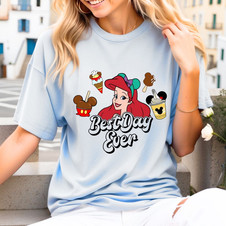 May include: Light blue t-shirt with a graphic of Ariel from The Little Mermaid, Mickey Mouse ears, and various Disney park treats. The text "Best Day Ever" is printed in a retro font.