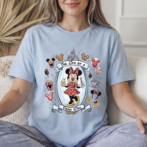 Op de afbeelding: Lichtblauw T-shirt met een cartoonontwerp van Minnie Mouse. Minnie draagt een rode polkadotjurk en houdt ijs en een traktatie vast. Het shirt heeft de tekst "The Life of a Park Girl" met Disney park iconen.