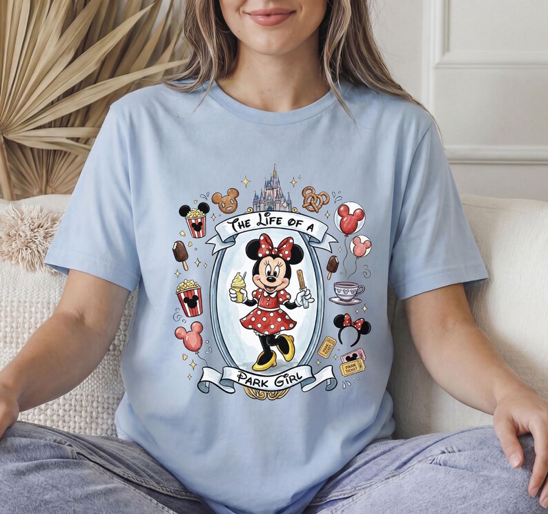 Op de afbeelding: Lichtblauw T-shirt met een cartoonontwerp van Minnie Mouse. Minnie draagt een rode polkadotjurk en houdt ijs en een traktatie vast. Het shirt heeft de tekst "The Life of a Park Girl" met Disney park iconen.