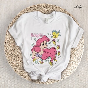 Peut inclure: T-shirt blanc avec un motif d'Ariel, la Petite Sir&egrave;ne, avec une queue rose et des poissons jaunes. Le texte "The Little Mermaid" est au-dessus d'Ariel.