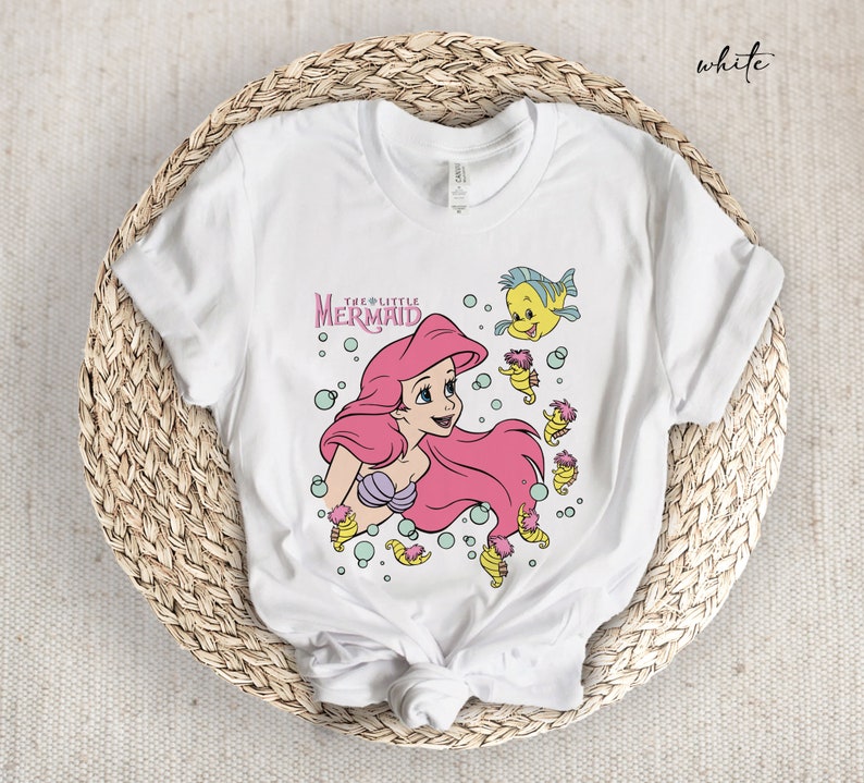 Peut inclure: T-shirt blanc avec un motif d'Ariel, la Petite Sir&egrave;ne, avec une queue rose et des poissons jaunes. Le texte "The Little Mermaid" est au-dessus d'Ariel.