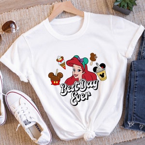 May include: White t-shirt featuring a graphic of Ariel from The Little Mermaid, surrounded by ice cream and treats. The text "Best Day Ever" is written in a playful font. The shirt is styled with white sneakers and sunglasses.