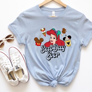 May include: Light blue t-shirt featuring a graphic of Ariel from The Little Mermaid, surrounded by cartoon treats. The text "Best Day Ever" is printed below the character. White sneakers and sunglasses are visible.