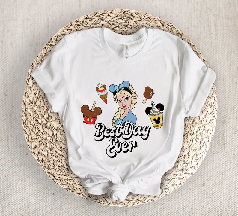 May include: White t-shirt with a graphic of a Disney princess with blonde hair and blue eyes. The princess is surrounded by Mickey Mouse ears and various Disney snacks. The text "Best Day Ever" is written in a whimsical font below the graphic.