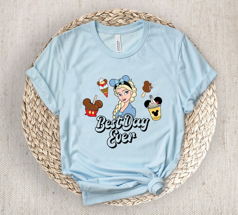 May include: Pale blue t-shirt with a cartoon of Elsa, ice cream, and the text "Best Day Ever". The shirt has rolled-up sleeves. The design includes various ice cream treats and a Mickey Mouse-shaped ice cream.