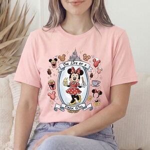 Op de afbeelding: Lichtroze T-shirt met een afbeelding van Minnie Mouse in een rode polkadotjurk met een ijsje. De tekst "The Life of a Park Girl" staat boven en onder de afbeelding, omringd door Disney-iconen.