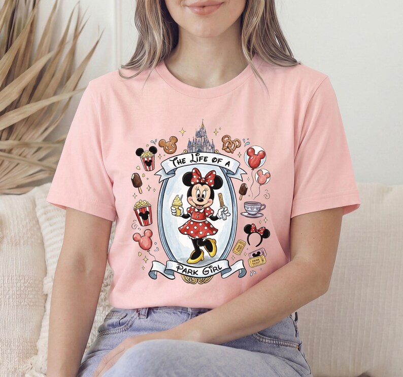 Op de afbeelding: Lichtroze T-shirt met een afbeelding van Minnie Mouse in een rode polkadotjurk met een ijsje. De tekst "The Life of a Park Girl" staat boven en onder de afbeelding, omringd door Disney-iconen.