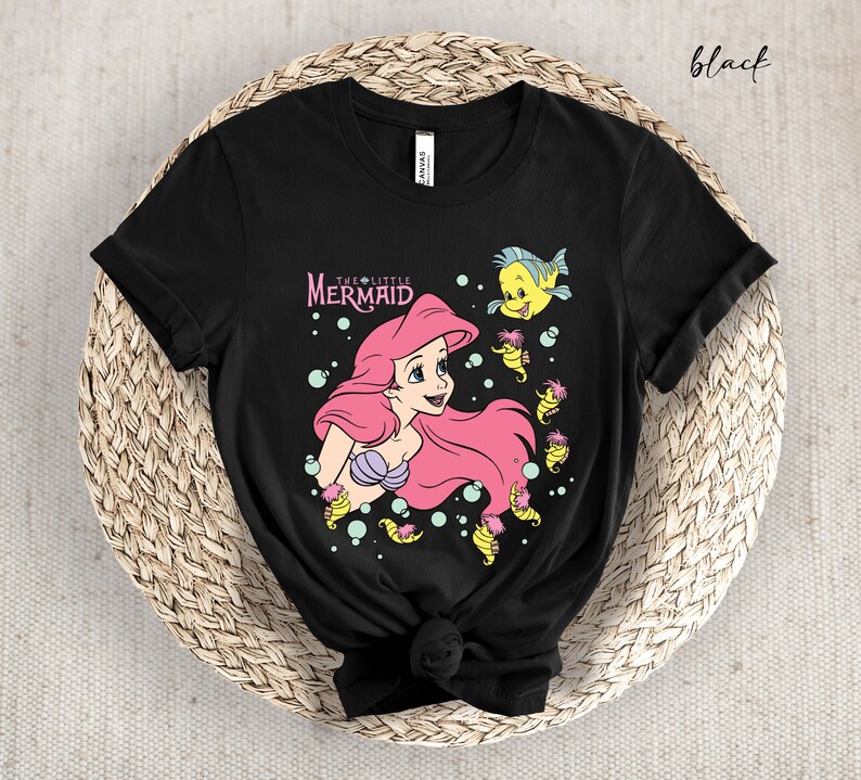 Peut inclure: T-shirt noir avec un motif d'Ariel de La Petite Sir&egrave;ne. Le motif repr&eacute;sente Ariel nageant avec un poisson jaune et des hippocampes. Le texte "The Little Mermaid" est imprim&eacute; au-dessus d'Ariel.