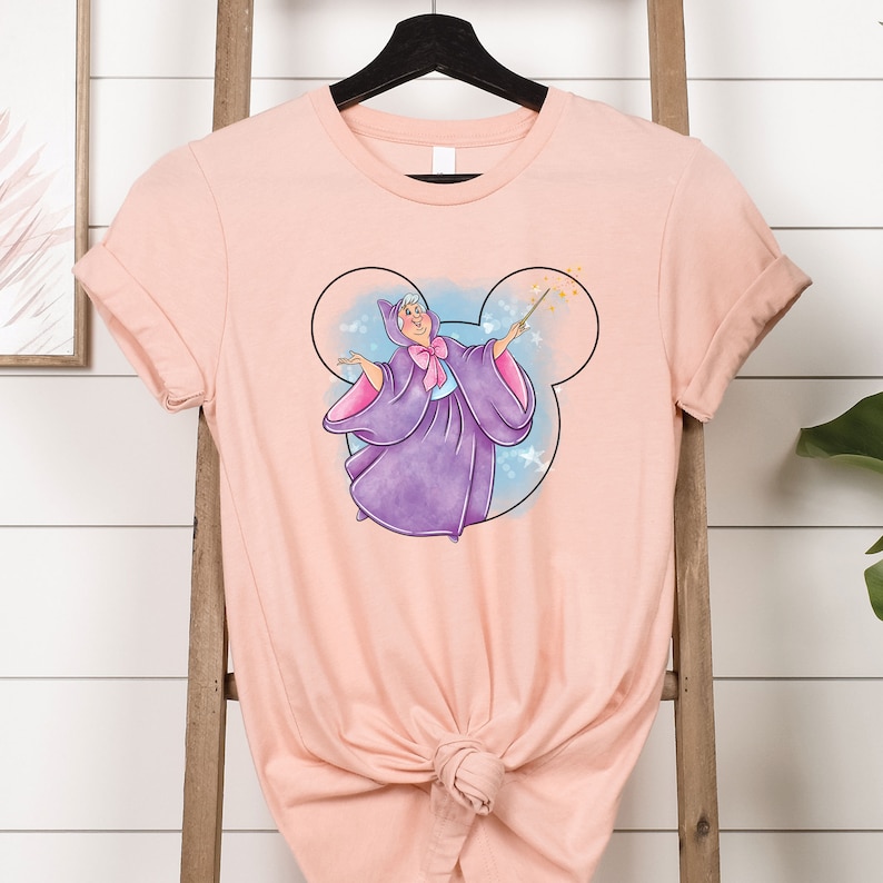 May include: A pink t-shirt with a graphic of Tinkerbell, the fairy from Disney's Peter Pan, flying inside a Mickey Mouse ear silhouette. The graphic is in shades of purple, blue, and gold.