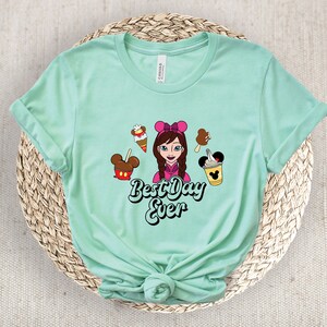 May include: Pale green t-shirt with a cartoon graphic of a girl with a pink bow, ice cream, and the text "Best Day Ever". The shirt is made of a soft material.