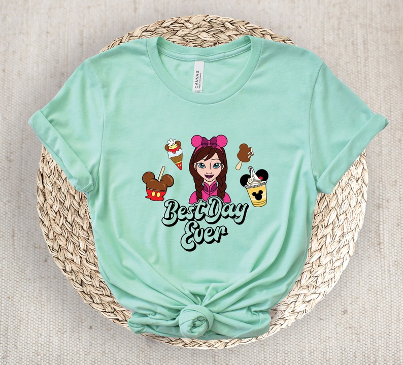 May include: Pale green t-shirt with a cartoon graphic of a girl with a pink bow, ice cream, and the text "Best Day Ever". The shirt is made of a soft material.