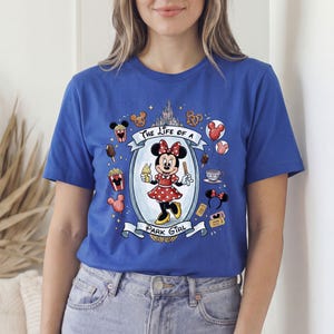Op de afbeelding: Koningsblauw T-shirt met een Minnie Mouse-afbeelding. De afbeelding toont Minnie Mouse in een rode polkadotjurk, de tekst "The Life of a Park Girl" en park-thema-iconen zoals popcorn en ijs.