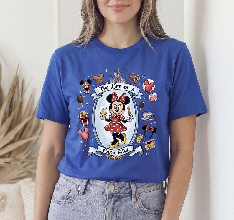 Op de afbeelding: Koningsblauw T-shirt met een Minnie Mouse-afbeelding. De afbeelding toont Minnie Mouse in een rode polkadotjurk, de tekst "The Life of a Park Girl" en park-thema-iconen zoals popcorn en ijs.