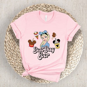 May include: A pale pink t-shirt with a cartoon illustration of Elsa, alongside ice cream treats. The text "Best Day Ever" is printed in black and white. The shirt is displayed on a woven basket.