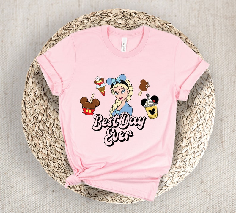 May include: A pale pink t-shirt with a cartoon illustration of Elsa, alongside ice cream treats. The text "Best Day Ever" is printed in black and white. The shirt is displayed on a woven basket.