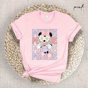 May include: Pink t-shirt with a graphic of Minnie Mouse wearing flowers over her eyes and the word "MINNIE" repeated in a retro font.