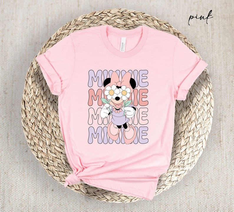 May include: Pink t-shirt with a graphic of Minnie Mouse wearing flowers over her eyes and the word "MINNIE" repeated in a retro font.
