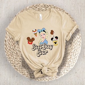 May include: A beige t-shirt with a cartoon of Elsa from Frozen, surrounded by ice cream treats and Mickey Mouse-shaped desserts. The text "Best Day Ever" is written in a retro font. The shirt is knotted at the bottom.