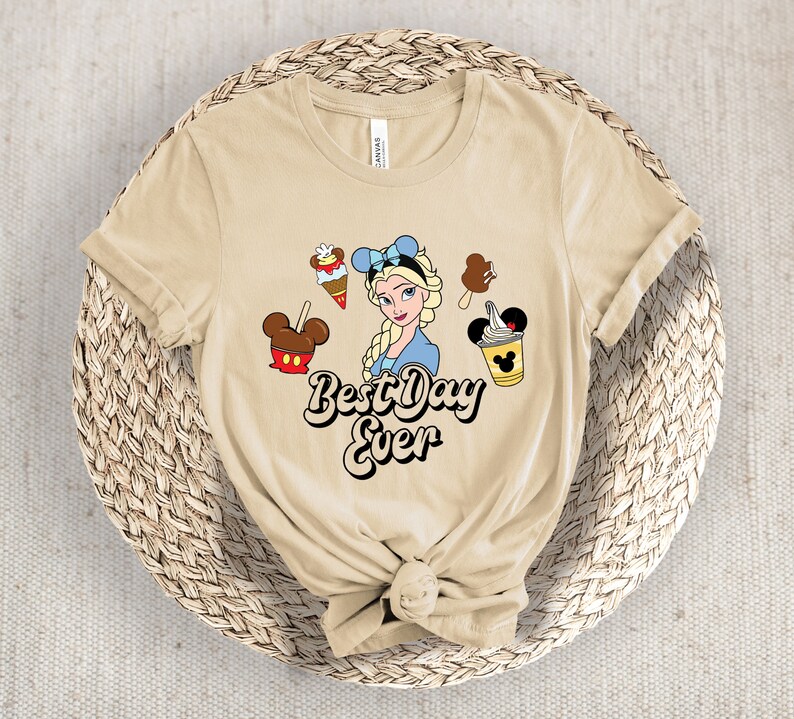 May include: A beige t-shirt with a cartoon of Elsa from Frozen, surrounded by ice cream treats and Mickey Mouse-shaped desserts. The text "Best Day Ever" is written in a retro font. The shirt is knotted at the bottom.