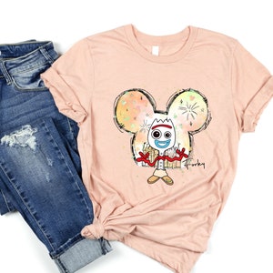 May include: A peach-colored t-shirt with a cartoon image of Forky from Toy Story. Forky is inside a Mickey Mouse head outline with a watercolor background. The text "Forky" is written below the image.