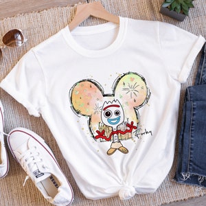 May include: White t-shirt with a cartoon drawing of Forky from Toy Story in a Mickey Mouse silhouette. The drawing is surrounded by a watercolor effect with fireworks and hearts. The text "Forky" is written below the drawing.