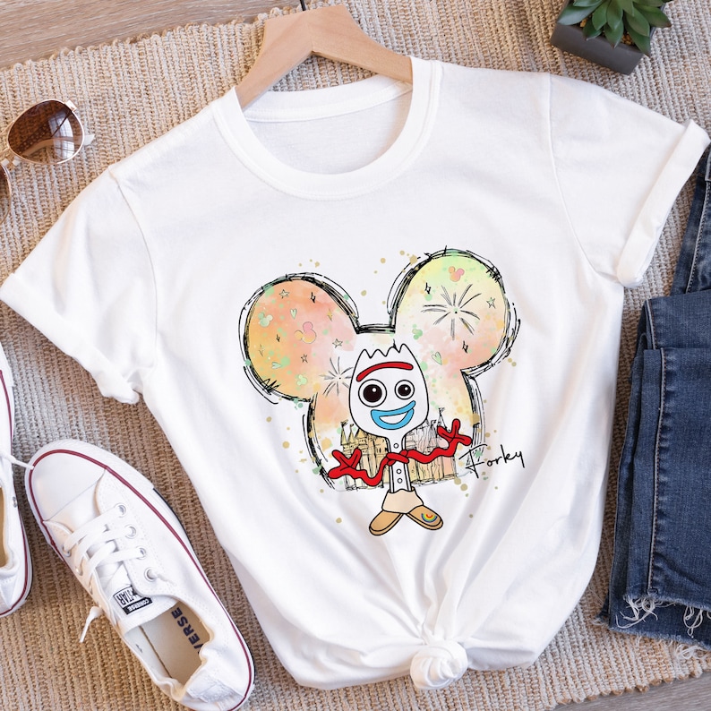 May include: White t-shirt with a cartoon drawing of Forky from Toy Story in a Mickey Mouse silhouette. The drawing is surrounded by a watercolor effect with fireworks and hearts. The text "Forky" is written below the drawing.