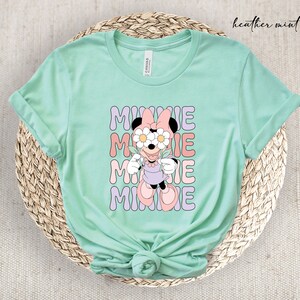 May include: A heather mint green t-shirt with a graphic of Minnie Mouse wearing flowers over her eyes. The text "MINNIE" is repeated four times in a retro font.