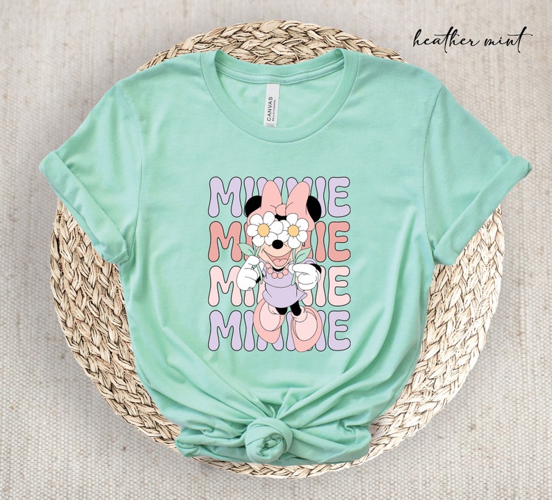 May include: A heather mint green t-shirt with a graphic of Minnie Mouse wearing flowers over her eyes. The text "MINNIE" is repeated four times in a retro font.