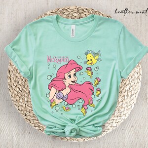 Peut inclure: Un t-shirt vert menthe clair avec un imprim&eacute; graphique d'Ariel de La Petite Sir&egrave;ne. Le graphique repr&eacute;sente Ariel nageant avec un poisson jaune et des hippocampes.