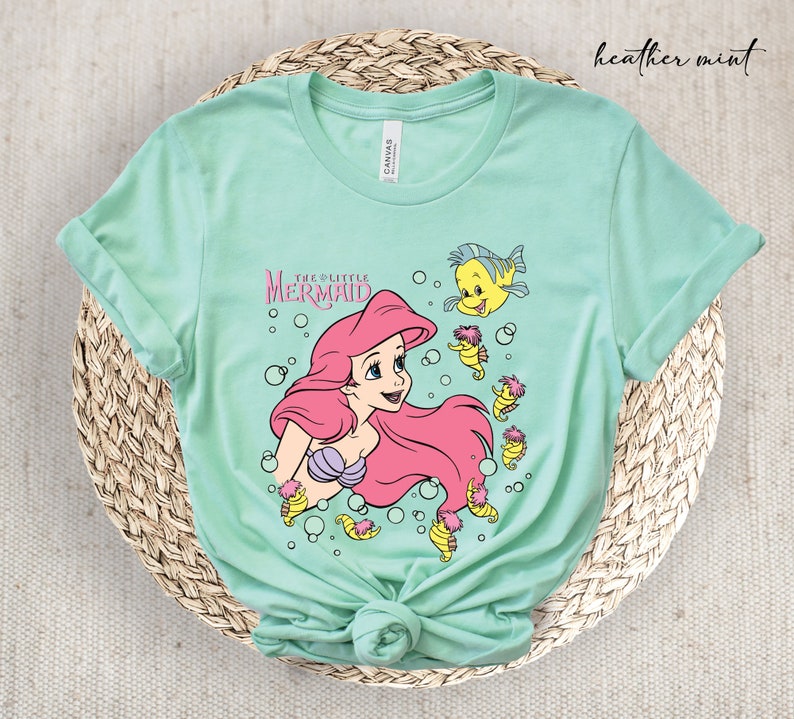 Peut inclure: Un t-shirt vert menthe clair avec un imprim&eacute; graphique d'Ariel de La Petite Sir&egrave;ne. Le graphique repr&eacute;sente Ariel nageant avec un poisson jaune et des hippocampes.