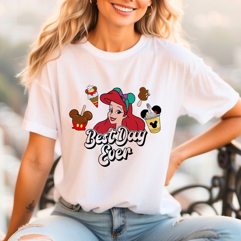 May include: White t-shirt featuring a graphic of Ariel from The Little Mermaid, surrounded by ice cream treats and a Mickey Mouse-shaped caramel apple. The text "Best Day Ever" is displayed below Ariel in a playful font.