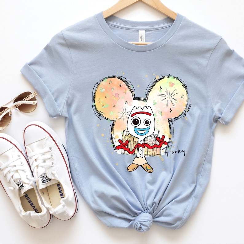 May include: Light blue t-shirt with a cartoon image of Forky from Toy Story. The image is inside a Mickey Mouse ear outline with a watercolor background. The text "Forky" is written below the image.