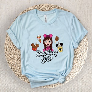 May include: Pale blue t-shirt featuring a cartoon girl, ice cream, and the text "Best Day Ever". The shirt is made of a soft material and has rolled-up sleeves. The graphic includes a Mickey Mouse ice cream bar.