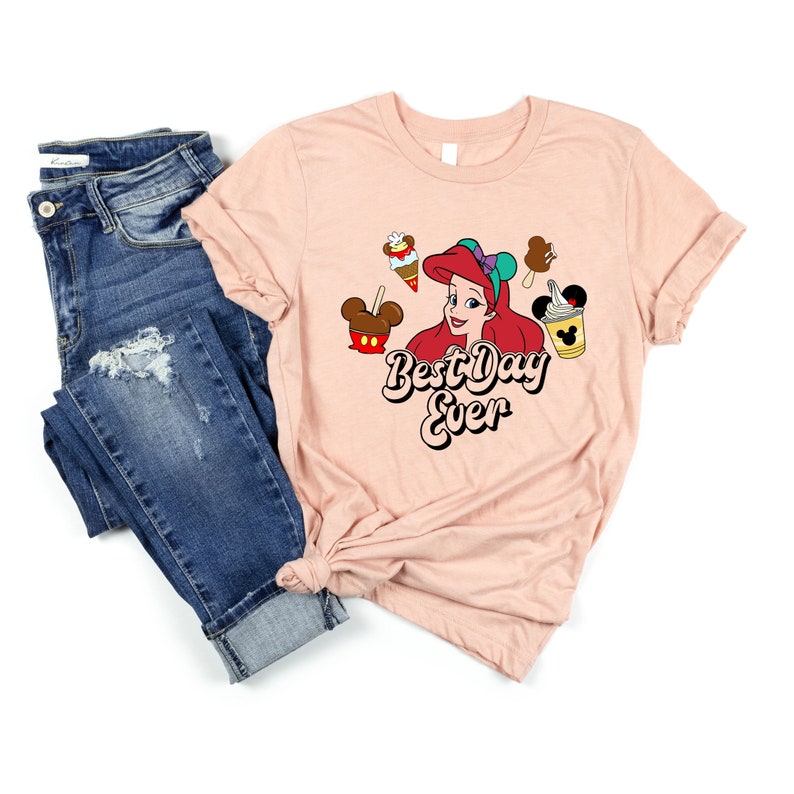 May include: A light pink t-shirt featuring a graphic of Ariel from The Little Mermaid, surrounded by Disney treats. The text "Best Day Ever" is displayed below Ariel. Blue denim jeans are folded next to the shirt.