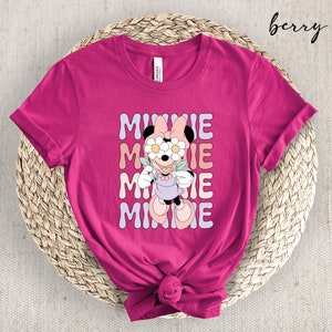 May include: A pink t-shirt with a graphic of Minnie Mouse wearing flowers over her eyes. The text "MINNIE" is repeated four times in a retro font.