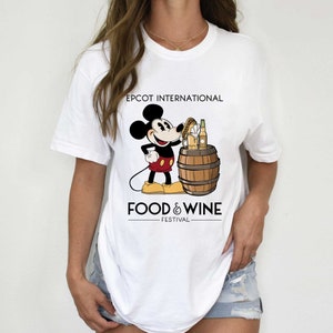 May include: White t-shirt featuring a black and white illustration of Mickey Mouse holding a beer and a pretzel. The text "EPCOT INTERNATIONAL FOOD & WINE FESTIVAL" is printed on the shirt.